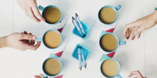 Six cups of tea and two pots with two pens in, taken from above with hands reaching for the cups