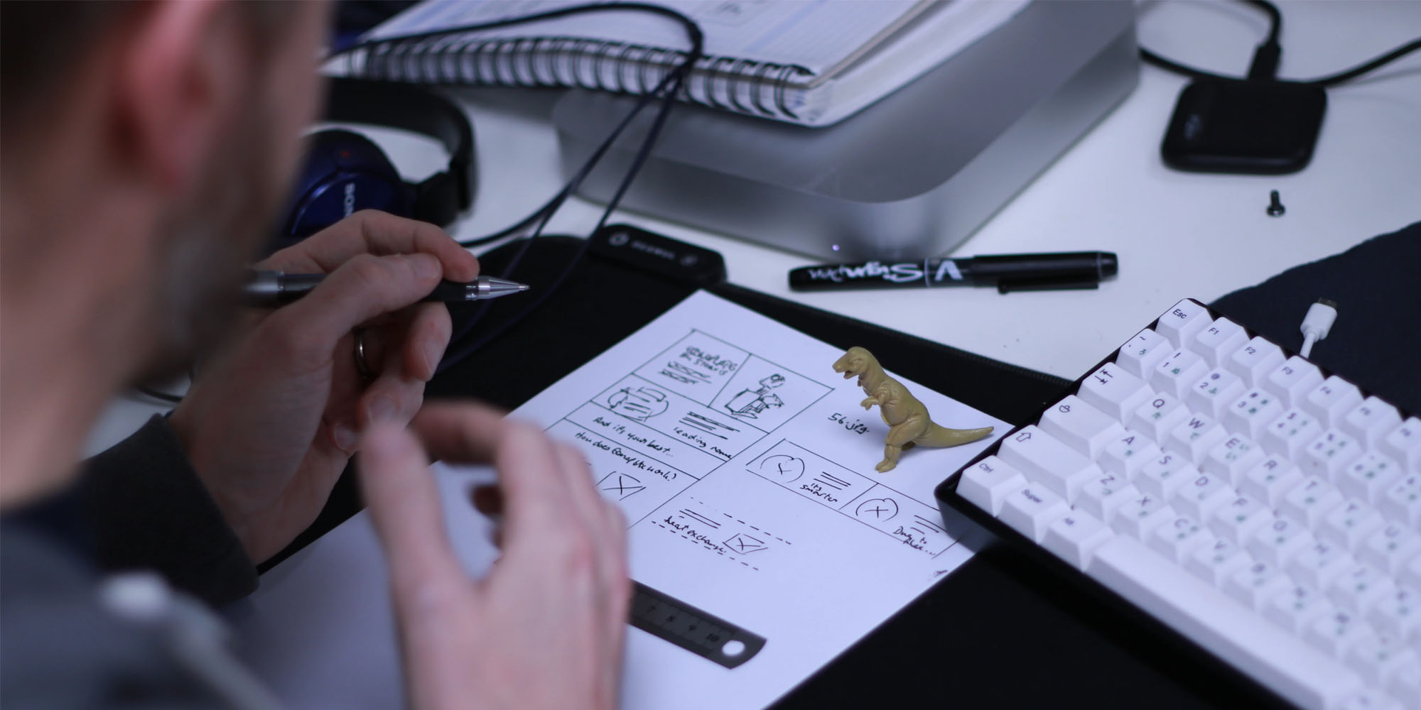 A person drawing layouts on a piece of paper, with a toy dinosaur at a desk