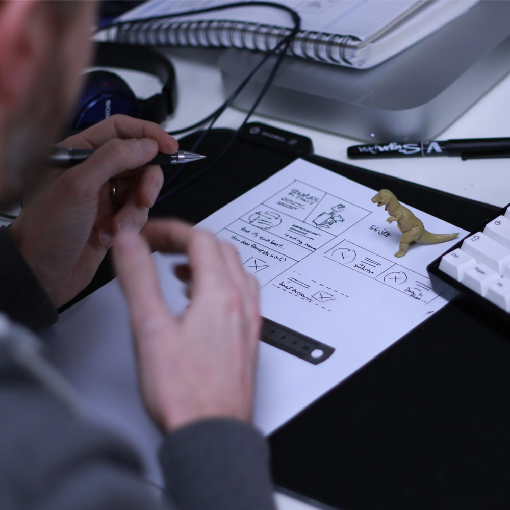 A person drawing layouts on a piece of paper, with a toy dinosaur at a desk