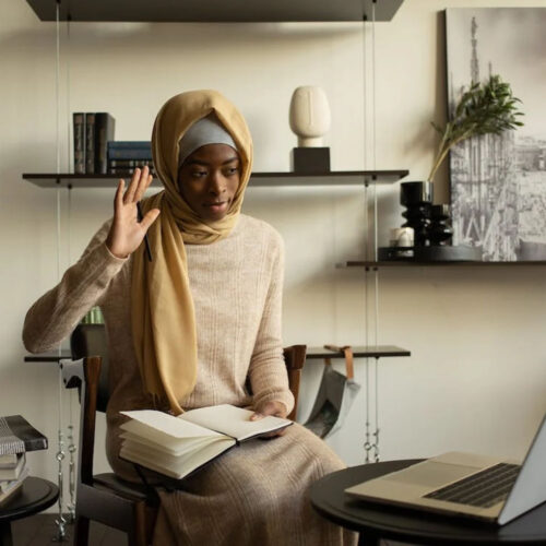 A woman wearing a beige headscarf waves at a laptop screen