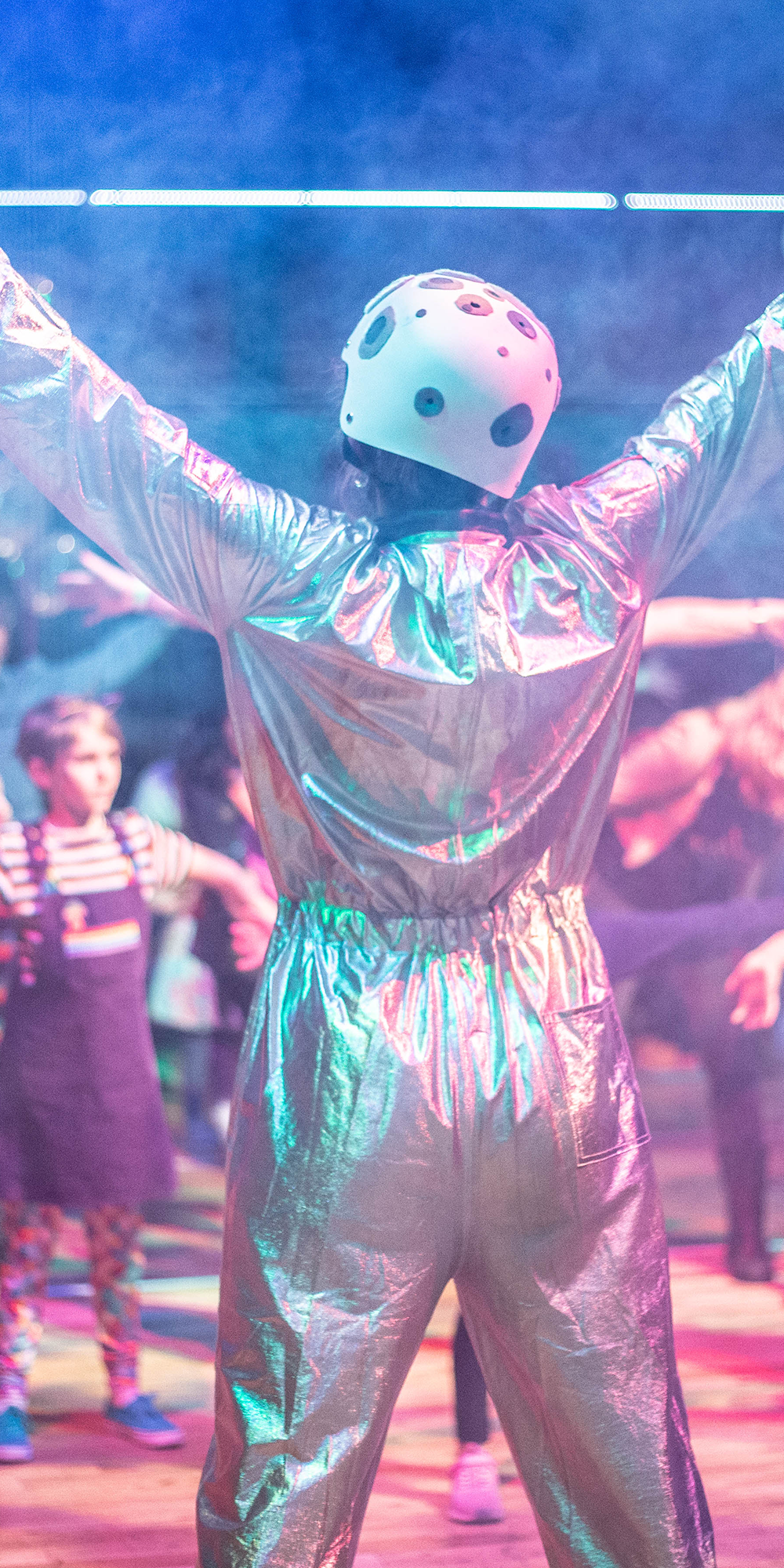 A person in a metallic silver jumpsuit and safety helmet, in front of a group of children in a smoky room