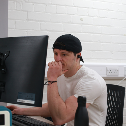 A man, Ash B, working on a computer, wearing a cap backwards