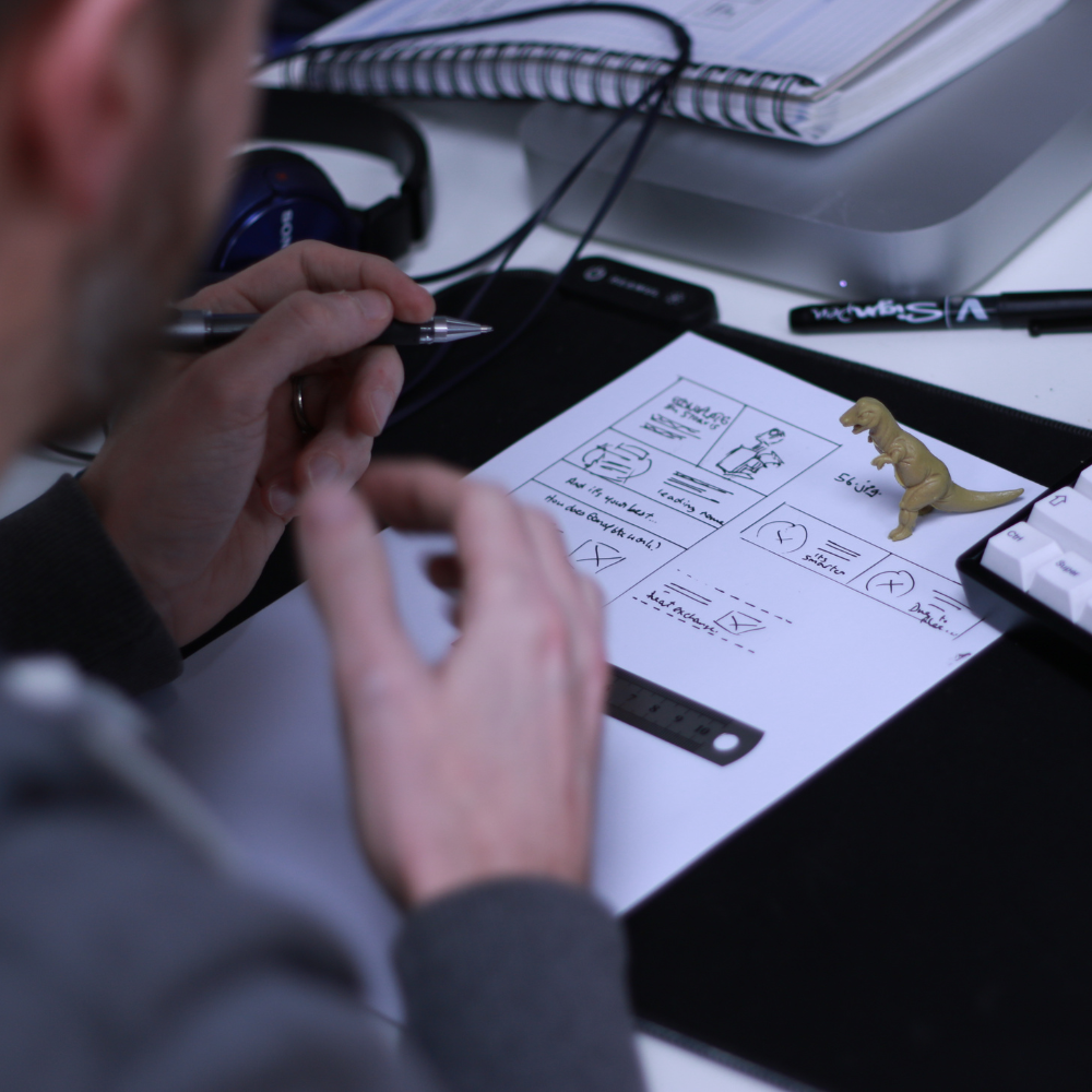 A paper on a desk with drawing and plans on, with a toy dinosaur on top