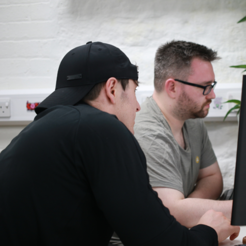 Two men looking at a computer screen, one is wearing a cap backwards